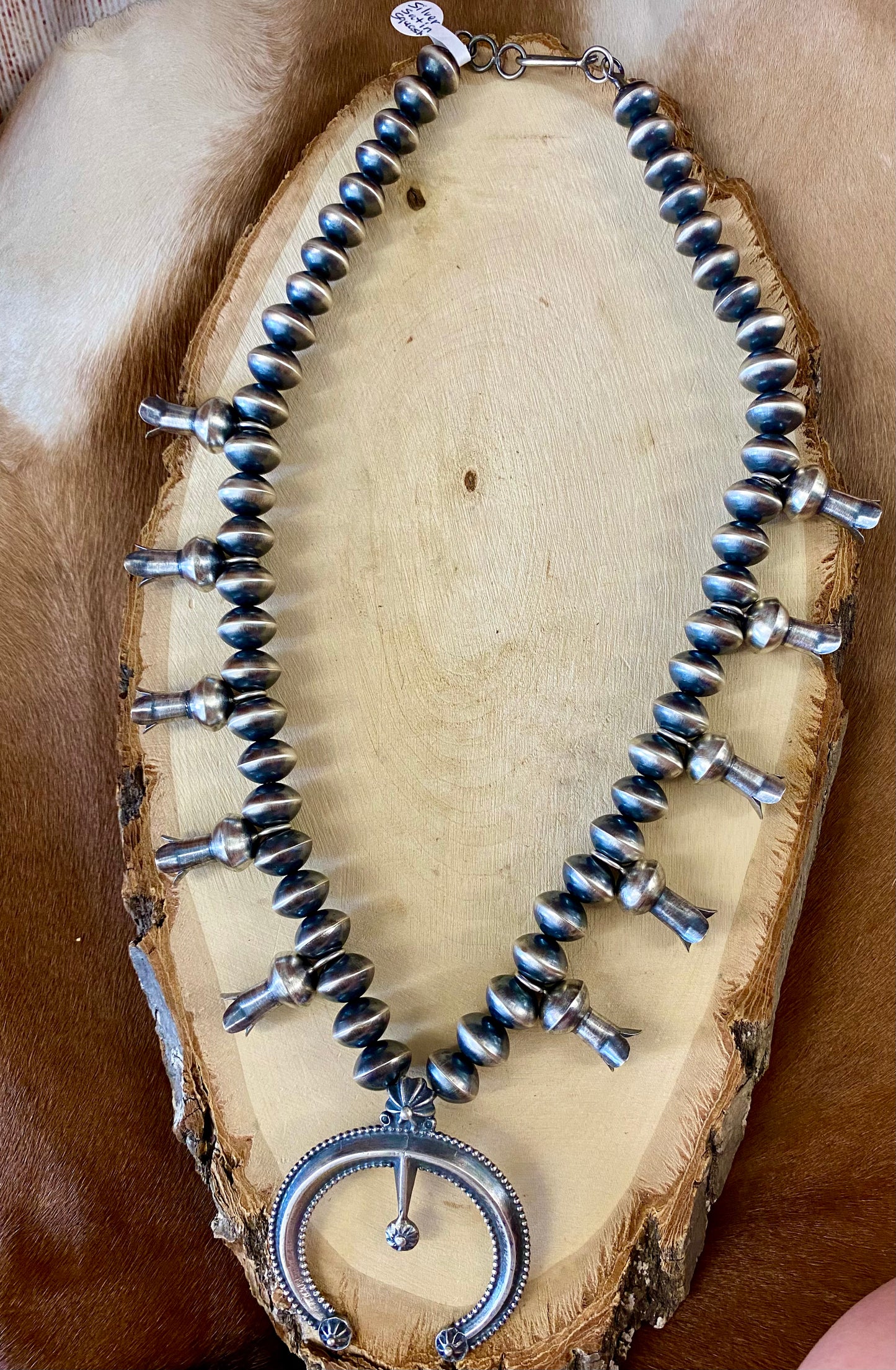 This sterling silver Silver Squash Blossom Necklace has a vintage feel. At the center, the Naja is 2-1/4" tall and 2-1/8" wide and is accented with 10 total flared blossoms 5 going up each side which are 1" inch long. These are strung on smooth, 12 mm Desert - Navajo Pearl strand. This necklace was given an old pawn satin buffing, and handcrafted by Native American artist and silversmith Presley Haley.