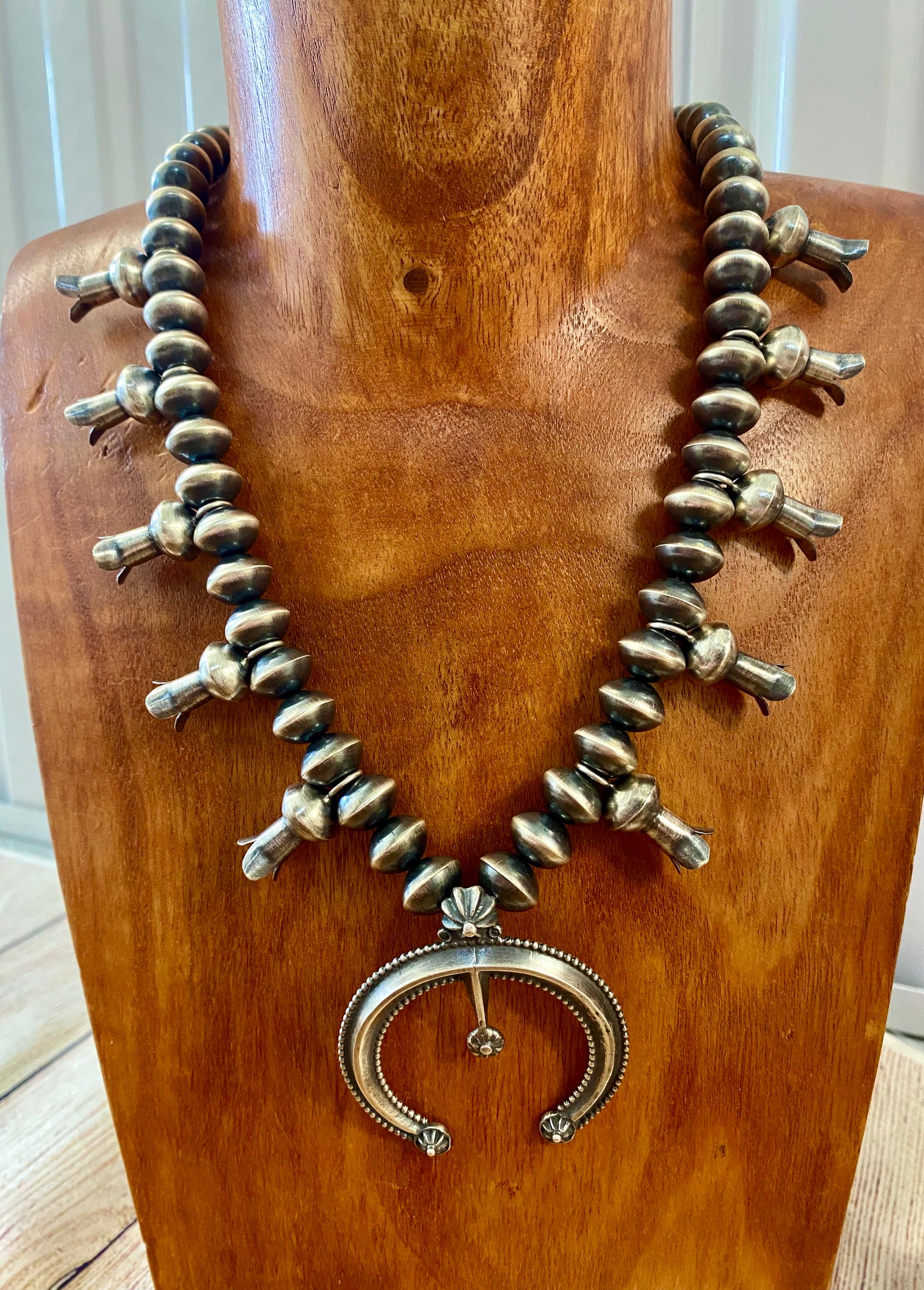 This sterling silver Silver Squash Blossom Necklace has a vintage feel. At the center, the Naja is 2-1/4" tall and 2-1/8" wide and is accented with 10 total flared blossoms 5 going up each side which are 1" inch long. These are strung on smooth, 12 mm Desert - Navajo Pearl strand. This necklace was given an old pawn satin buffing, and handcrafted by Native American artist and silversmith Presley Haley.