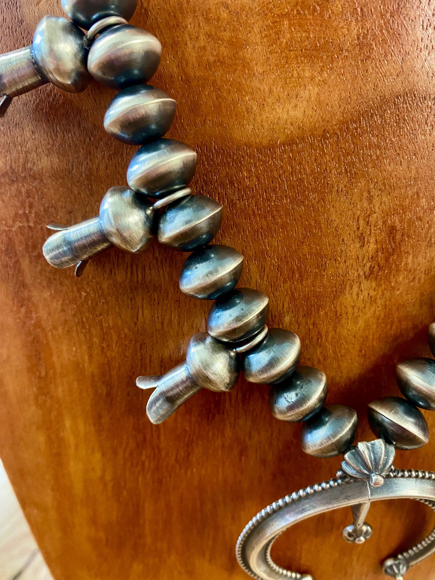 This sterling silver Silver Squash Blossom Necklace has a vintage feel. At the center, the Naja is 2-1/4" tall and 2-1/8" wide and is accented with 10 total flared blossoms 5 going up each side which are 1" inch long. These are strung on smooth, 12 mm Desert - Navajo Pearl strand. This necklace was given an old pawn satin buffing, and handcrafted by Native American artist and silversmith Presley Haley.