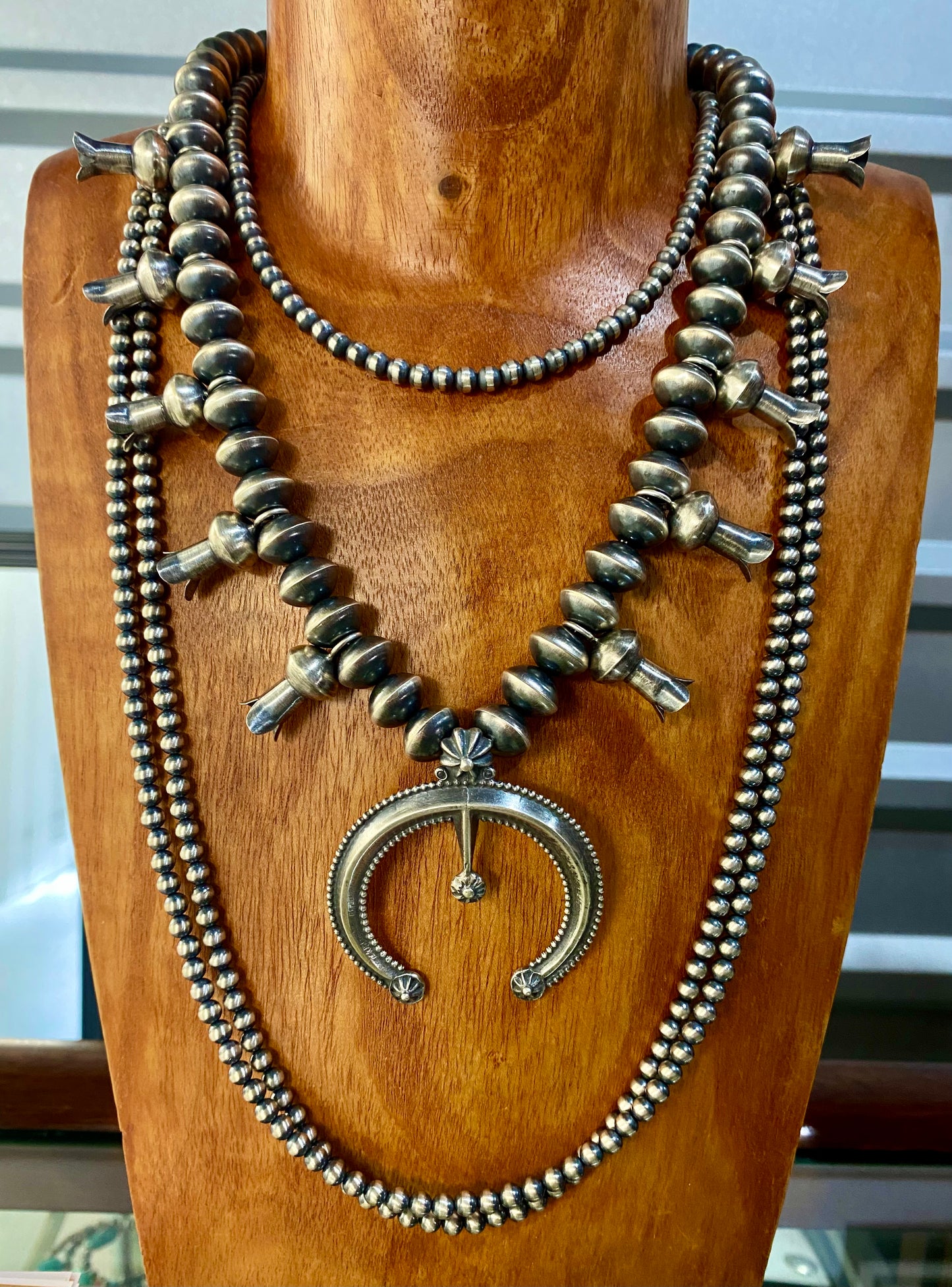 This sterling silver Silver Squash Blossom Necklace has a vintage feel. At the center, the Naja is 2-1/4" tall and 2-1/8" wide and is accented with 10 total flared blossoms 5 going up each side which are 1" inch long. These are strung on smooth, 12 mm Desert - Navajo Pearl strand. This necklace was given an old pawn satin buffing, and handcrafted by Native American artist and silversmith Presley Haley.