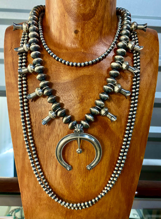This sterling silver Silver Squash Blossom Necklace has a vintage feel. At the center, the Naja is 2-1/4" tall and 2-1/8" wide and is accented with 10 total flared blossoms 5 going up each side which are 1" inch long. These are strung on smooth, 12 mm Desert - Navajo Pearl strand. This necklace was given an old pawn satin buffing, and handcrafted by Native American artist and silversmith Presley Haley.